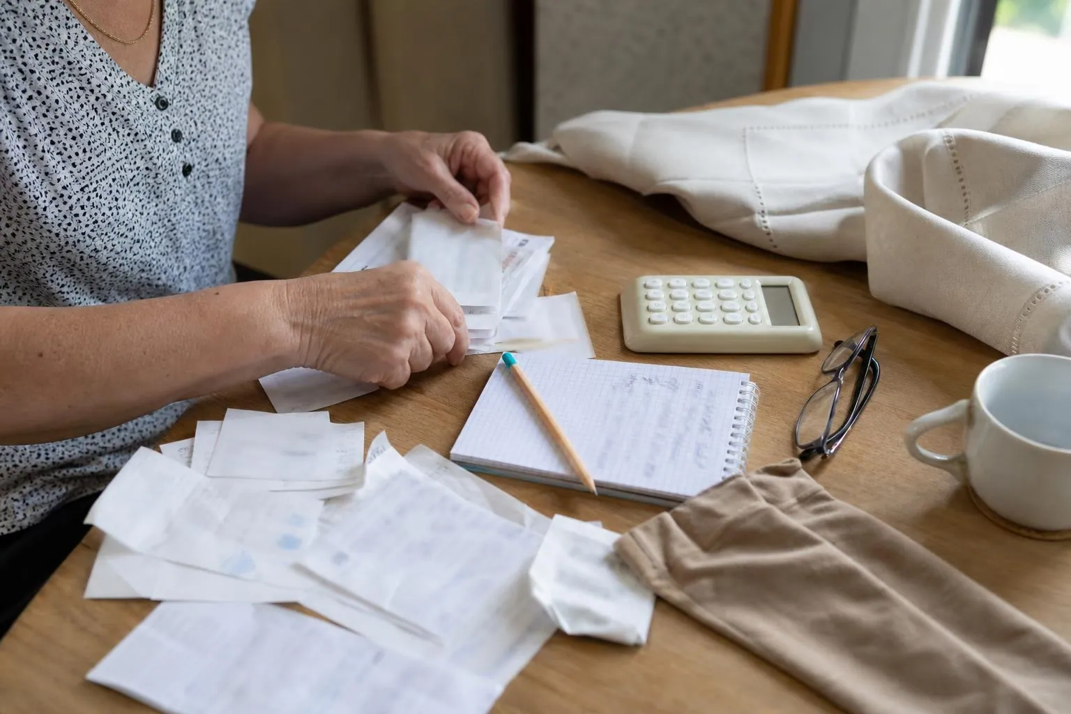 Gros plan sur une table où une femme aligne des factures pliées, deux bas de compression usagés et un carnet quadrillé annoté au crayon